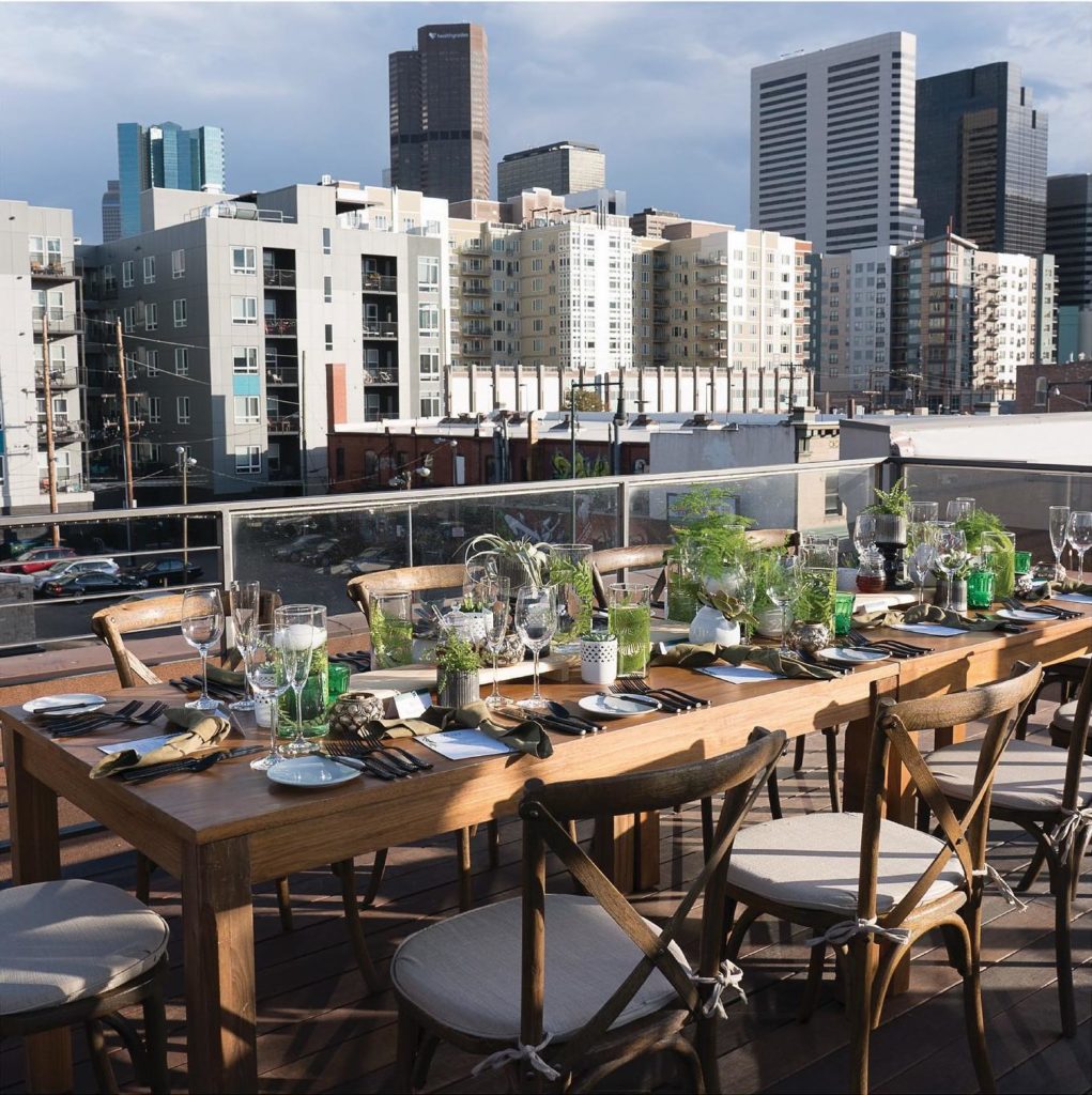 Rustic dining table with chairs on a roof top with black silverware, green cups, and greenery details. In the background are lots of buildings.