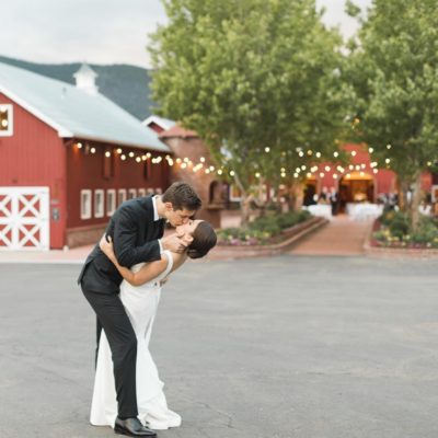 A groom and bride passionately kissing in from of a red barn venue with trees and string lights