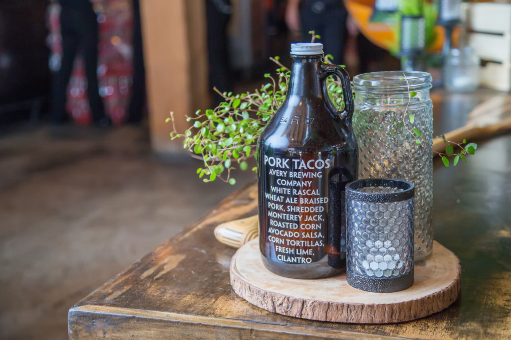 A Growler bottle with white text in all caps that reads "Pork Tacos" in large font "avery brewing company white rascal wheat ale braised pork, shredded monterey jack, roasted corn, avocado salsa, corn tortillas, fresh lime, cilantro" on a wood circle on a wooden table with a tall glass mason jar with tecture and a black votive with hexagon pattern
