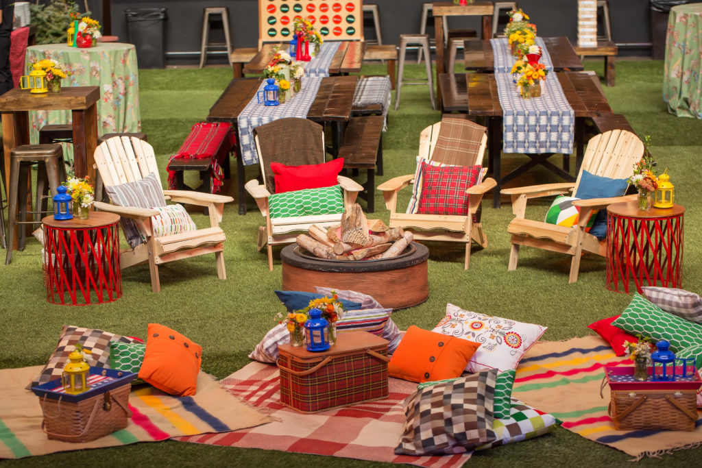 Wooden outdoor chairs on fake grass with colorful blankets and pillows laid out. Picnic baskets and lanterns laid out.