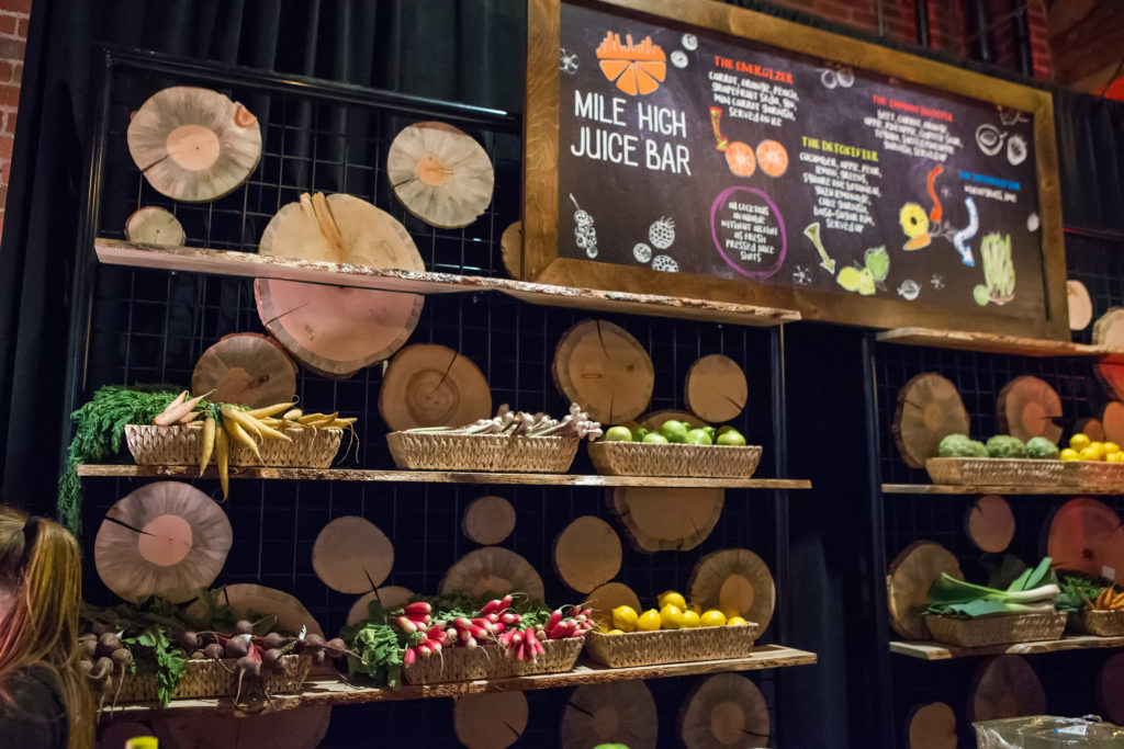 A juice bar with baskets full of full vegetables and fruits with a beetle kill wall. Above the walls is a wooden and chalkboard sign that reads 'Mile High Juice Bar'