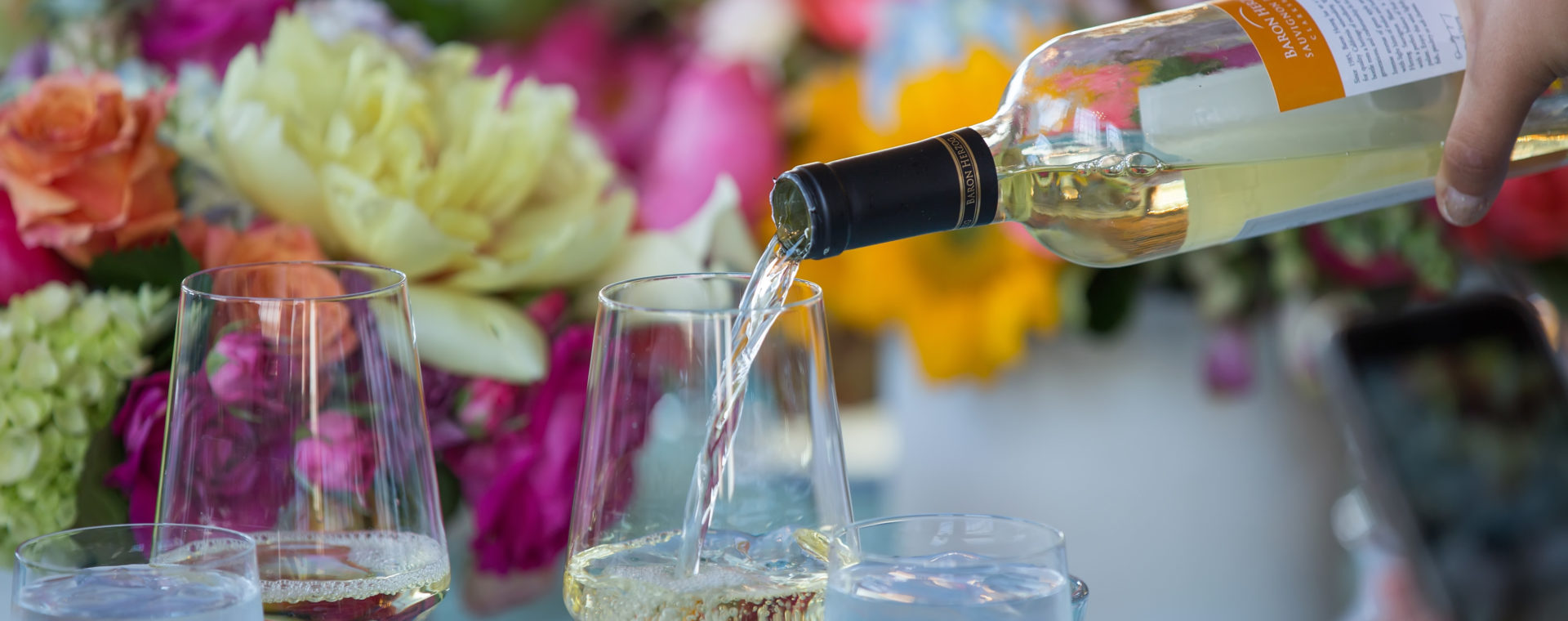 A hand pouring white wine into a wine glass with beautiful bright florals in the background