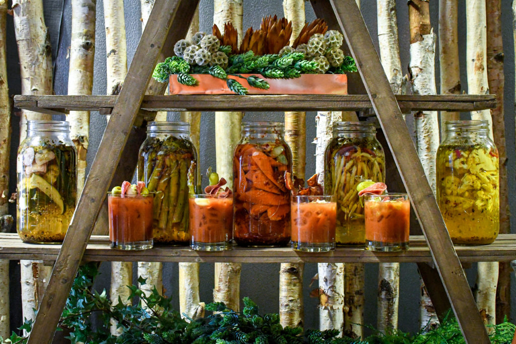 Four bloody mary cocktails on an wooden A-frame with mason jars of pickled vegetables behind it