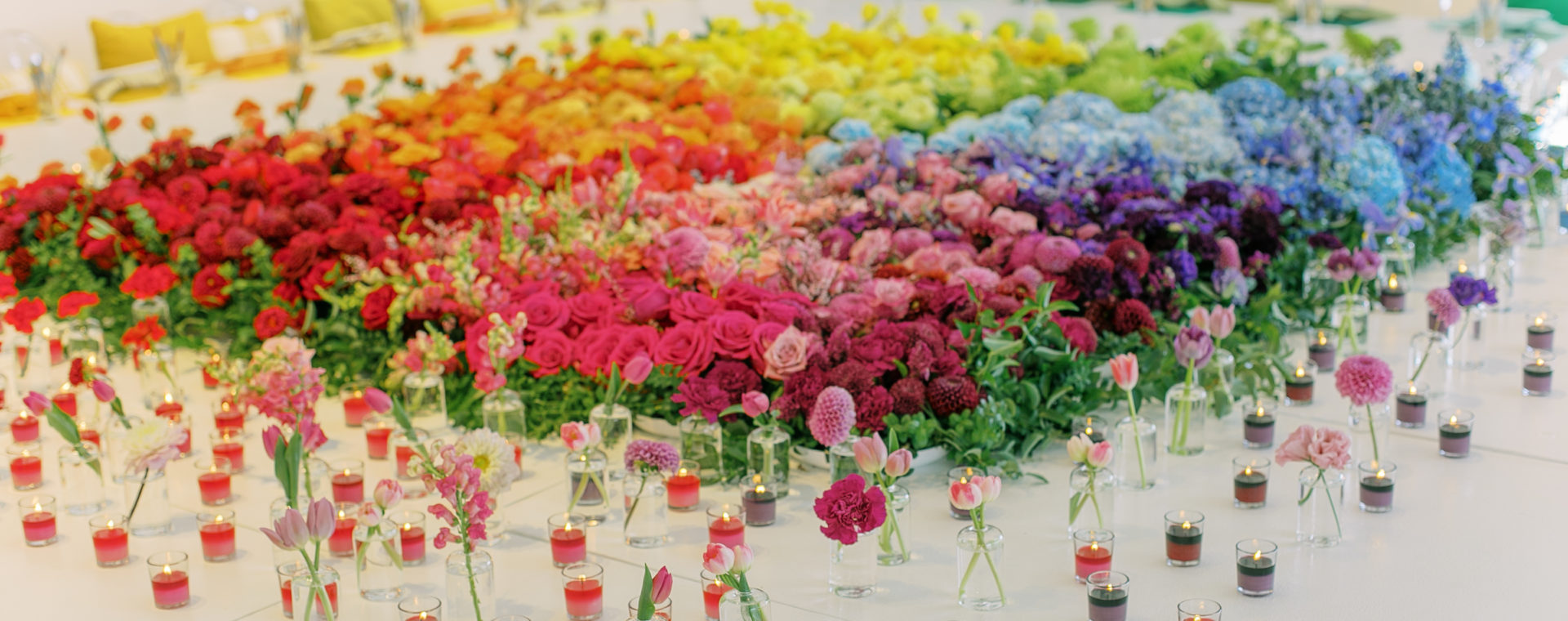 A giant white table covered with flowers in the center in a rainbow ombre and scattered single glass bottles with a single flower and votives with colorful candles