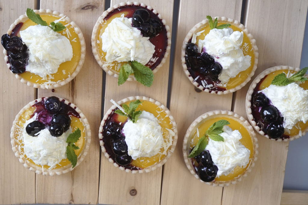 Mini tarts with lemon curd, blueberries, whipped cream, shaved white chocolate and mint on light wooden planks