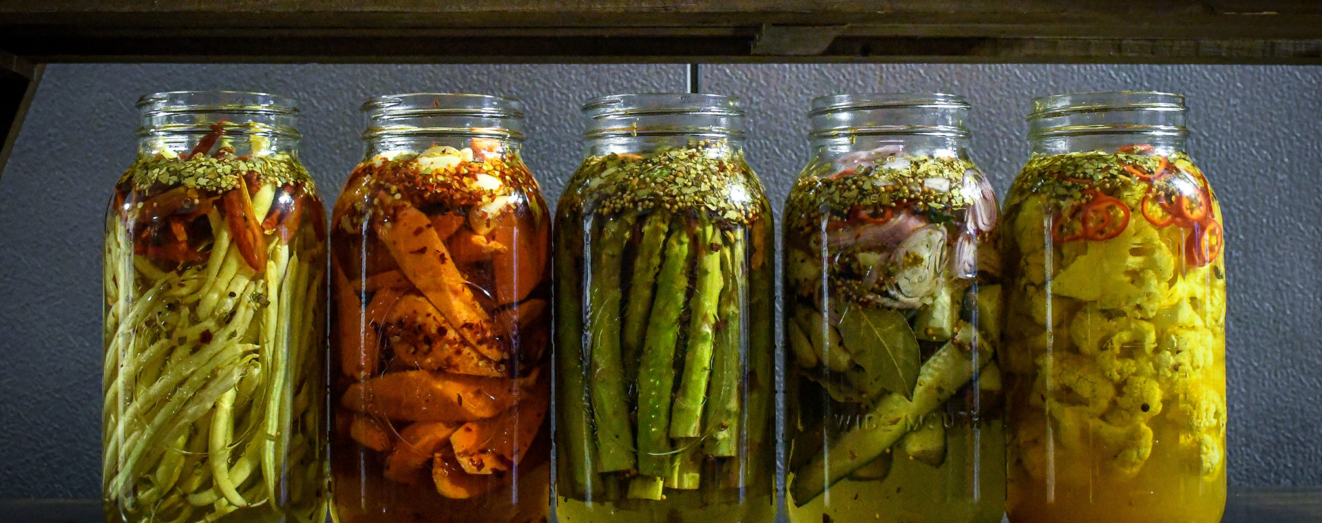 5 large mason jars with different pickled vegetables. From left to right, peppers, carrots, asparagus, cucumbers and cauliflower.