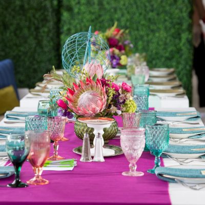 Close up of a colorful set table with a purple runner, blue and pink goblet glasses, and colorful floral centerpieces