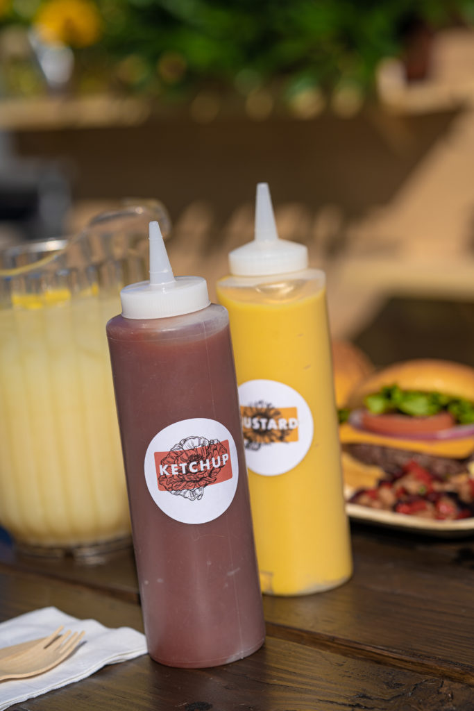 custom ketchup and mustard bottles on a wooden tables. Behind the bottles is a pitcher of cold lemonade and cheeseburgers.