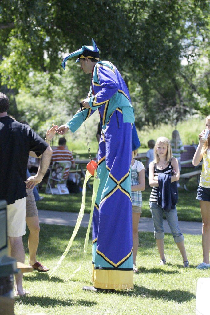 A man dresses as a jester in a blue outfit is on stilts outsider interacting with people