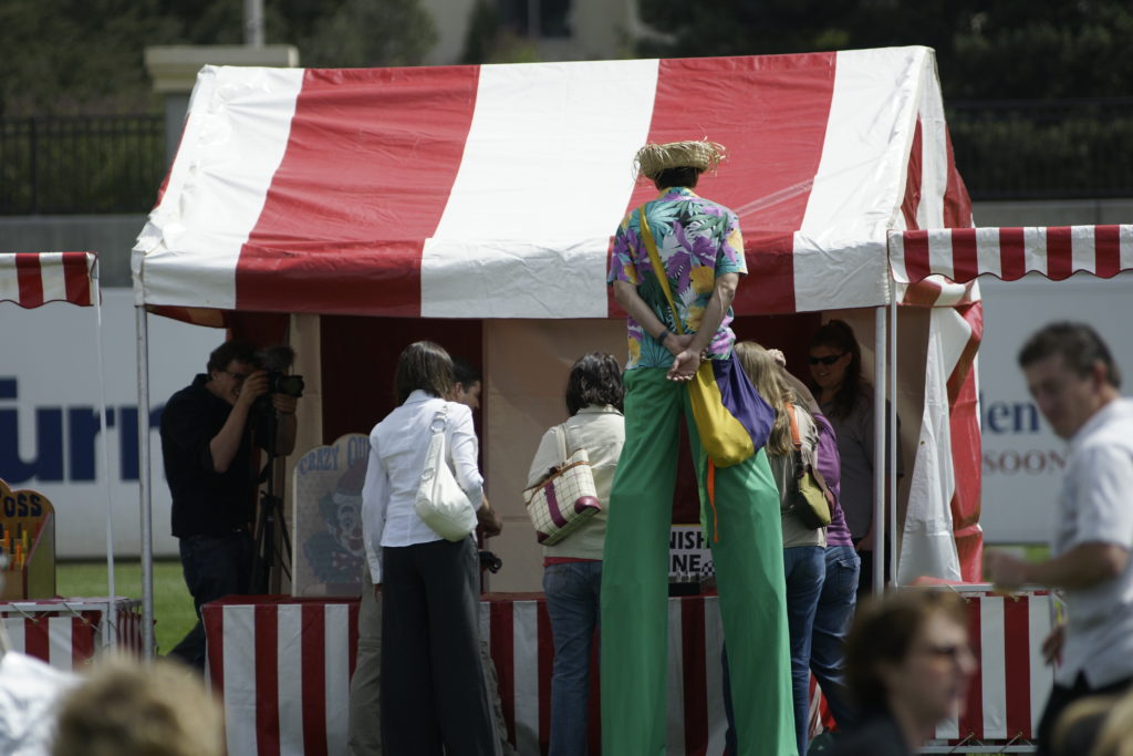 A man on stilts is standing behind a carnival game with red and white stipes