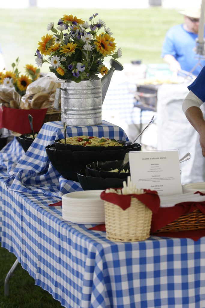 A buffet table with a blue gingham tablecloth with picnic food onto top and yellow and white floral arrangements