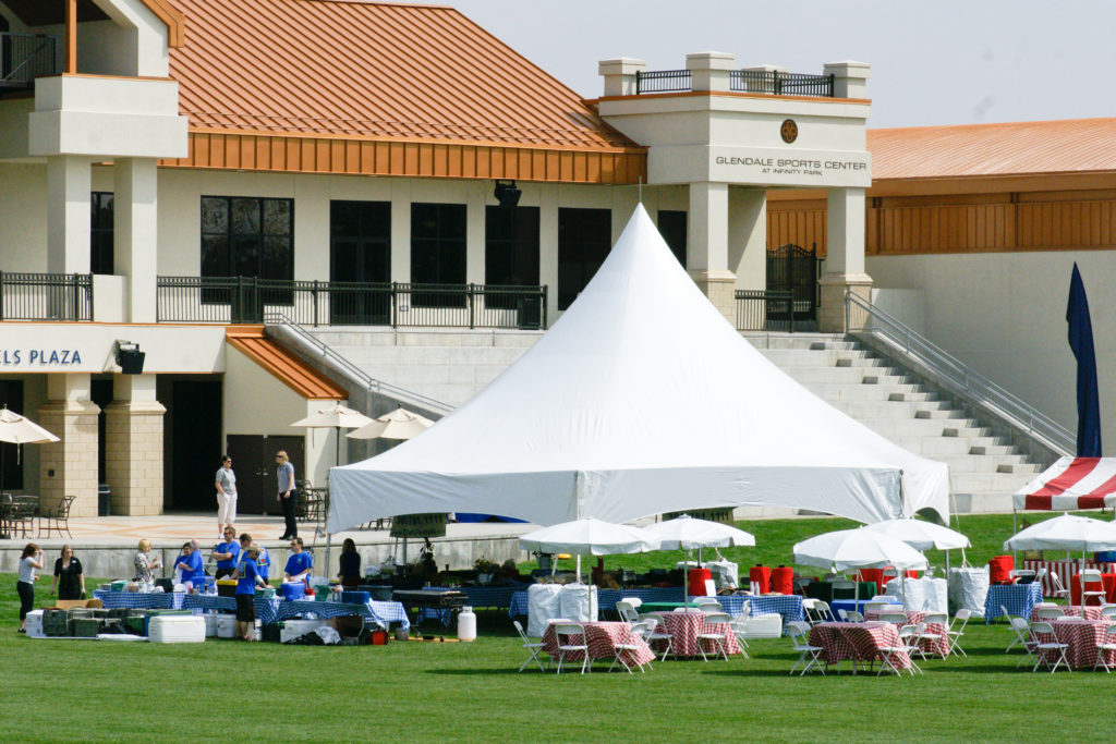 A large white tent outside of the glendale sports center set up for a picnic with round tables with gingham table linen, and white umbrellas
