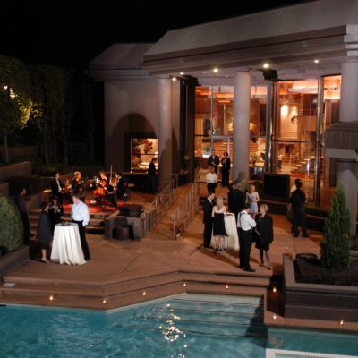 Outside a large house-like venue, with people in cocktail attire around the patio by the pool. A small group of musicians are playing to the upper left of the patio