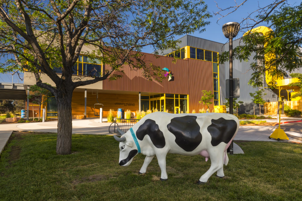 A venue made up of two buildings. One brown and one grey with yellow accents. In front of the venue is a lawn with a tree and a cow statue.