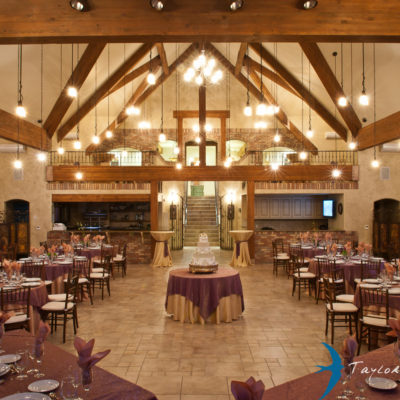 Inside a mountain chateau venue set up for a wedding reception with a circle table in the center with a large multiple story cake. Surrounding it are circle and rectangle tables with a burgundy tablecloths.