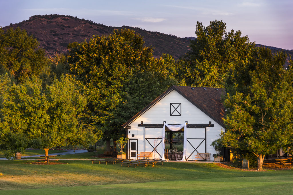 A large white barn surrounded by large trees. Outside the large barn doors are benches set up for a wedding ceremony