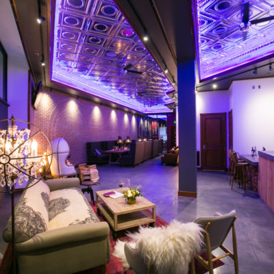 Inside the Denver Speakeasy, their is lounge furniture set up with a neon purple wall that is glowing on the silver tiles of the ceiling