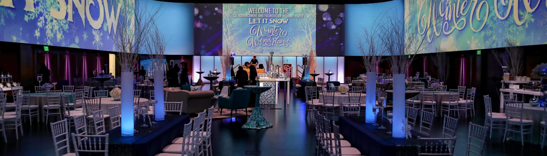 Inside an open venue set up for a winter holiday party with dimmed lighting, screens showing snowflakes and a variety of tables and chairs set up for a dinner