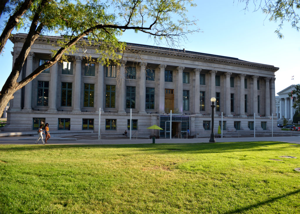 A full view of a wide building with a two people waking by on the sidewalk on a sunny day with a clear blue sky