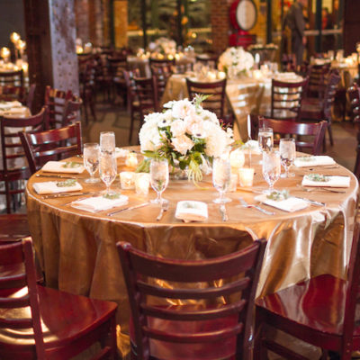 A circle table with a gold table cloth and mahogany chairs surrounding it. On the table are a white floral arrangement.