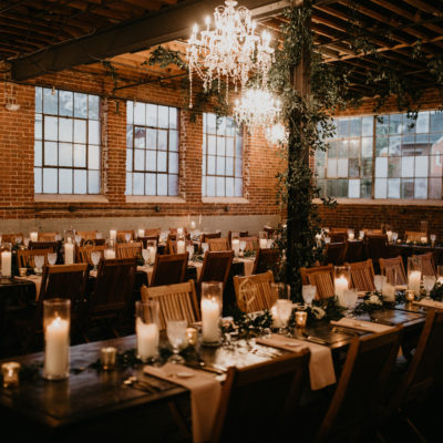 A red brick room set up for a wedding reception with long wooden tables and wooden chairs with tall candles, votives and greenery on each table. The room is dimly lit and the only light is coming from the candles and large crystal chandlers