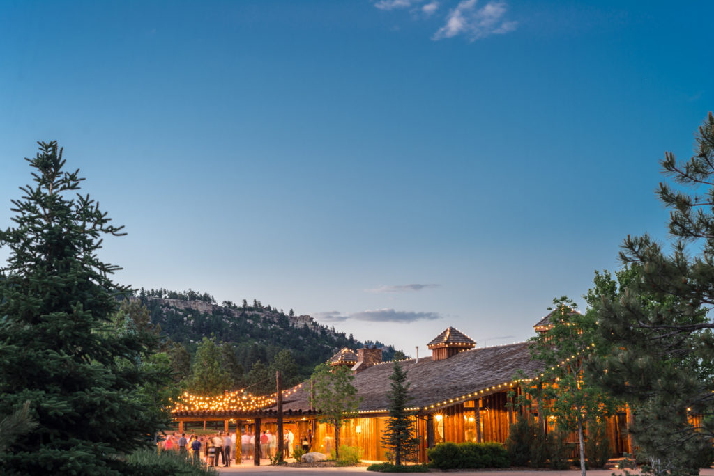 People outside a ranch venue among pine trees and mountains with the sunsetting in the background