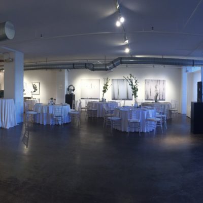 Inside Walker Fine Arts with cement flour, large open windows, white tables and silver chairs and paintings in surrounding