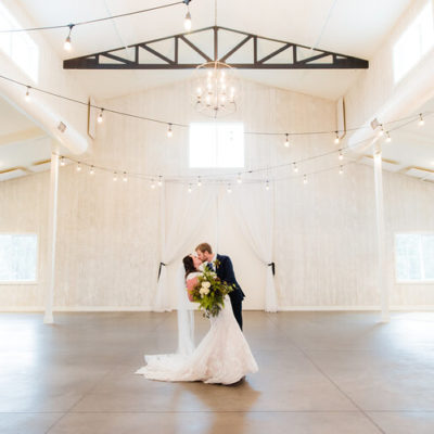 A bride and groom kissing in the middle of inside a white barn with lights hanging from the ceiling