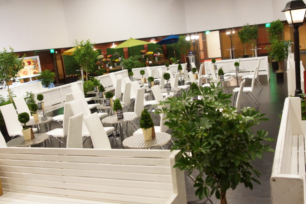 Inside Infinity Park, set up as an outdoor restaurant/park with white benches, trees in planters and small silver tables with white chairs and a small plant on each table