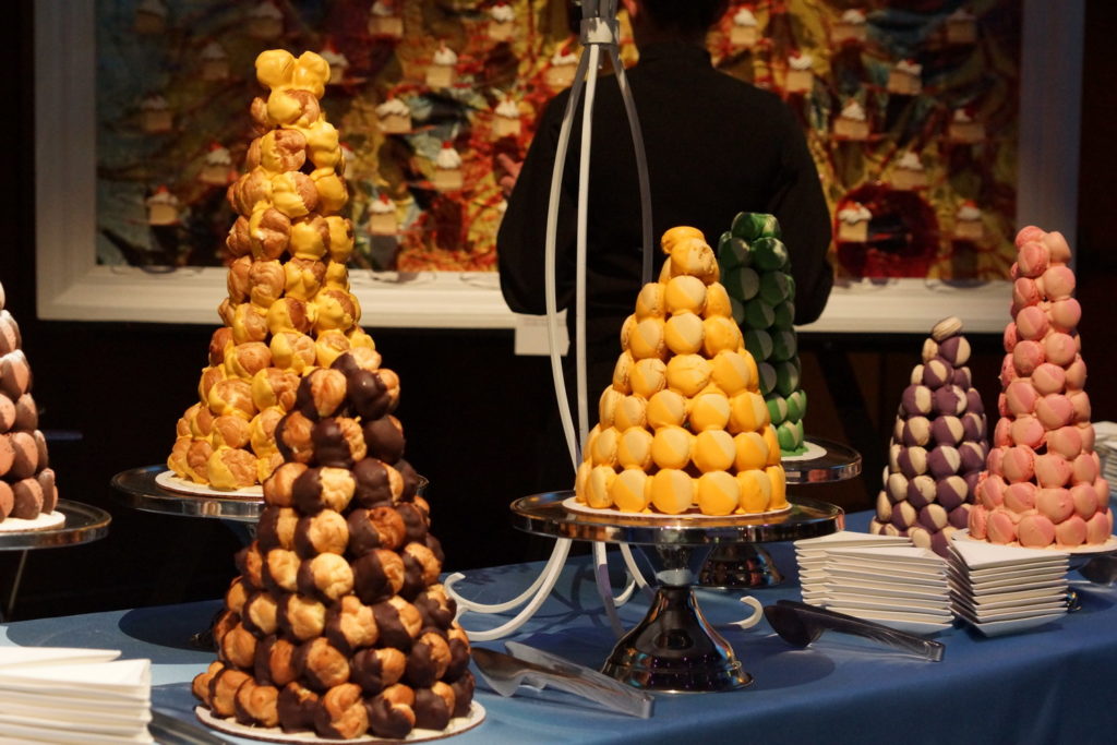 Colorful macaron towers and croquembouches sit on a buffet table with a blue linen and small triangle white plates