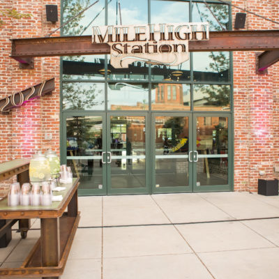 Right outside the doors of mile high station. THe sign that reads "Mile High Station" is made from metal with 2 large set of doors underneath and the building is made from red brick. Right in front of the building is a metal table with infused water.