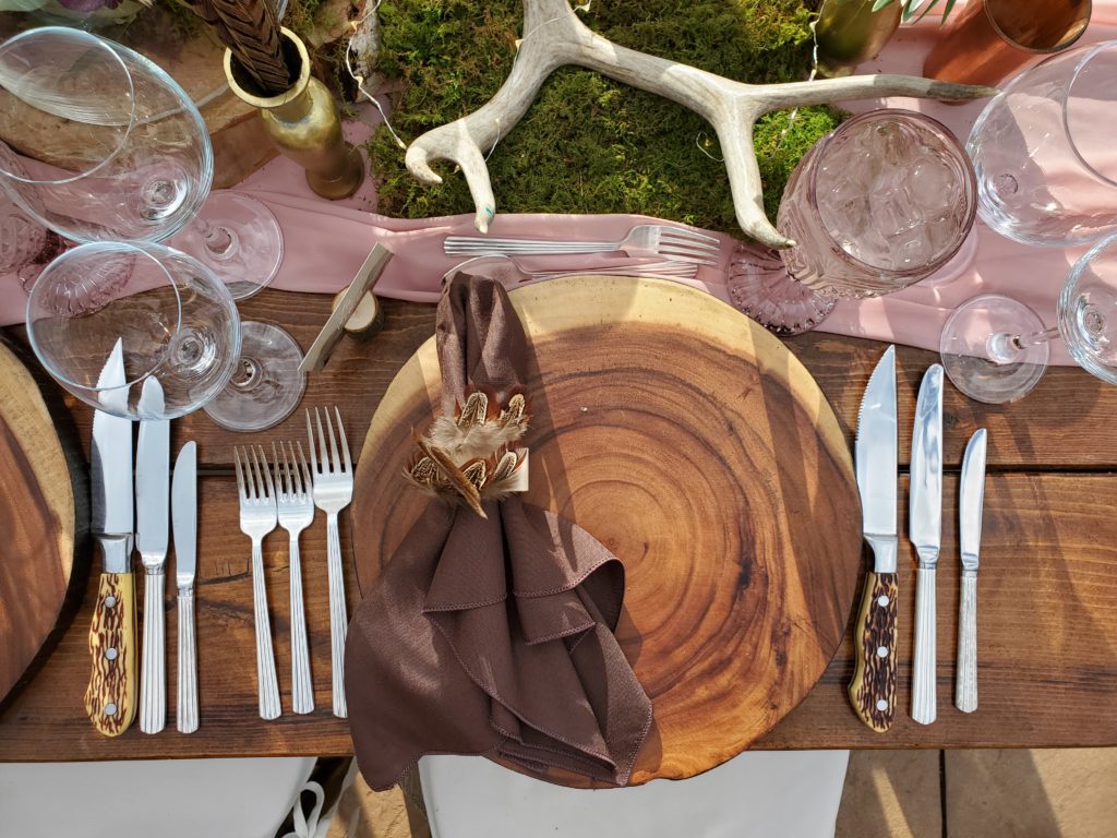 A close up of a place seating on a wooden table made from a large wood placemat with a brown napkin on top with with a feather napkin ring. At the place setting are 3 forks to the left, above is a dessert spoon and fork and to the right of the setting are three different knives consisting of a steak knife, dinner knife and butter knife. In the center of the table is a pink table runner with moss as decoration