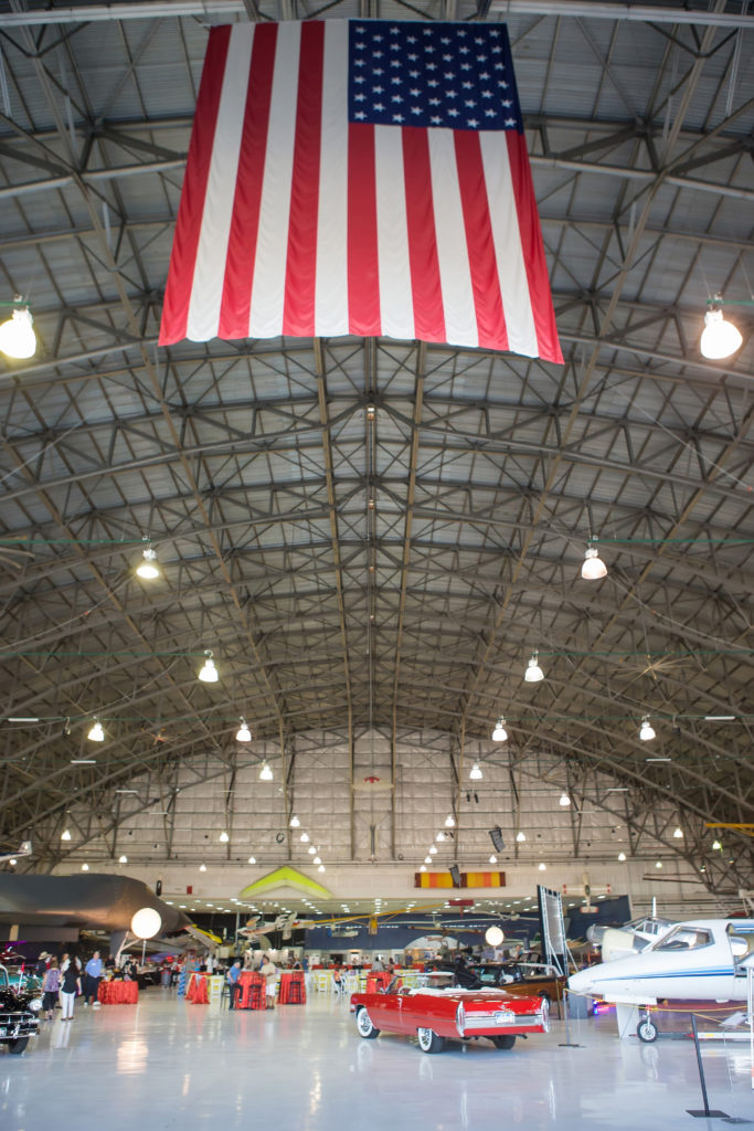 A focus on the ceiling at Wings over the Rockies, a large American Flag hands from the ceiling. On the floor are antique cars amount the planes and it is set up with lots of tall tables for sitting.