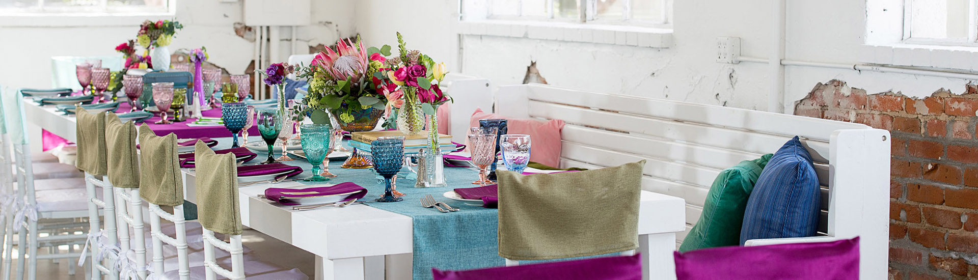 White table with white benches with pink, green and blue pillows, and white chairs with a white sea cover and olive green set cover. Blue linen table runner with a deep purple napkins and blue, teal and pink goblet glasses. In the center are flower arrangements with books underneath it.