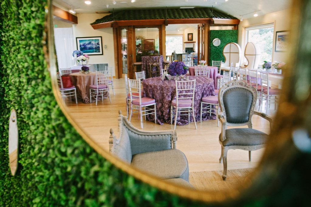 A mirror reflection attached to a hedge wall. In the mirror is there are different types of tables. In the center, is a circle pate with a purple table linen with rosettes on it. On the left are a small circle table with a plain purple linen. To the far right is a white rectangle table. On the tables are florals are purple, pink, light blue and white. In the front are a grey chair and grey couch.