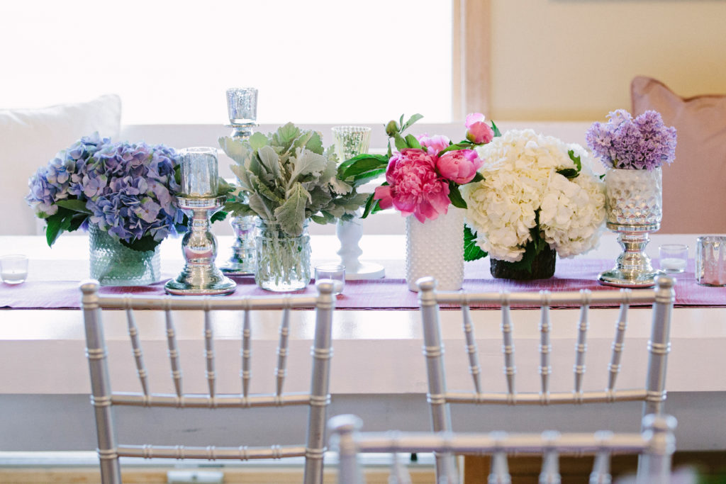 Close up of a white table. In the center is a purple runner. In the left is a blue hydrangeas, to the right is a bunch of greenery, to the right of that are pink peonies in a white dotted mason jar, to the right of it are white hydrangeas, to the right of that are small lilac flowers in a silver save. Surrounding the flowers are silver and glass votives. In front of the table are 2 silver chairs.