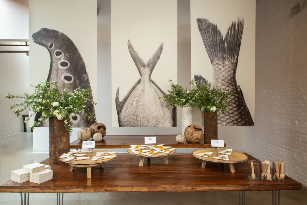 A wooden table with three small triangle plates with different fish on each plate. Behind is another wooden table with wooden balls and florals and above the table are 3 large black and white prints of different fish tails