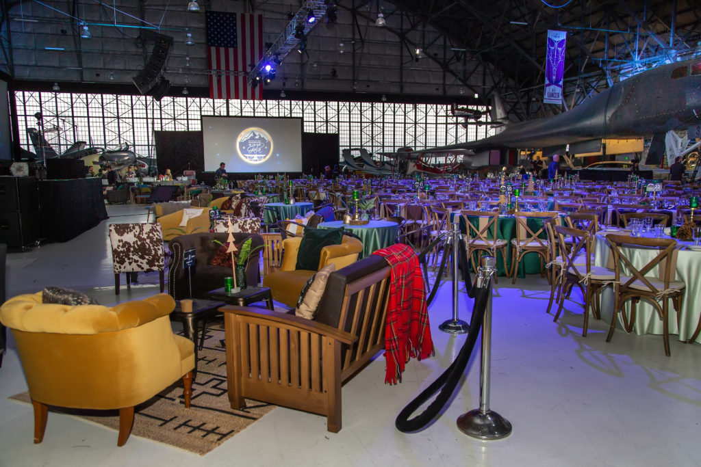 Airplaines in the background of he hanger, with a large American flag hanging down in the background. Underneath the flag is a large screen that has a logo of mountains and a plane with the text "Spreading Wings Gala". In the middle are a bunch of round tables with different shades of green linen with wooden chairs. To the left, are some additional seating as couches and comfy arm chairs in different shades of brown, yellow, animal print.