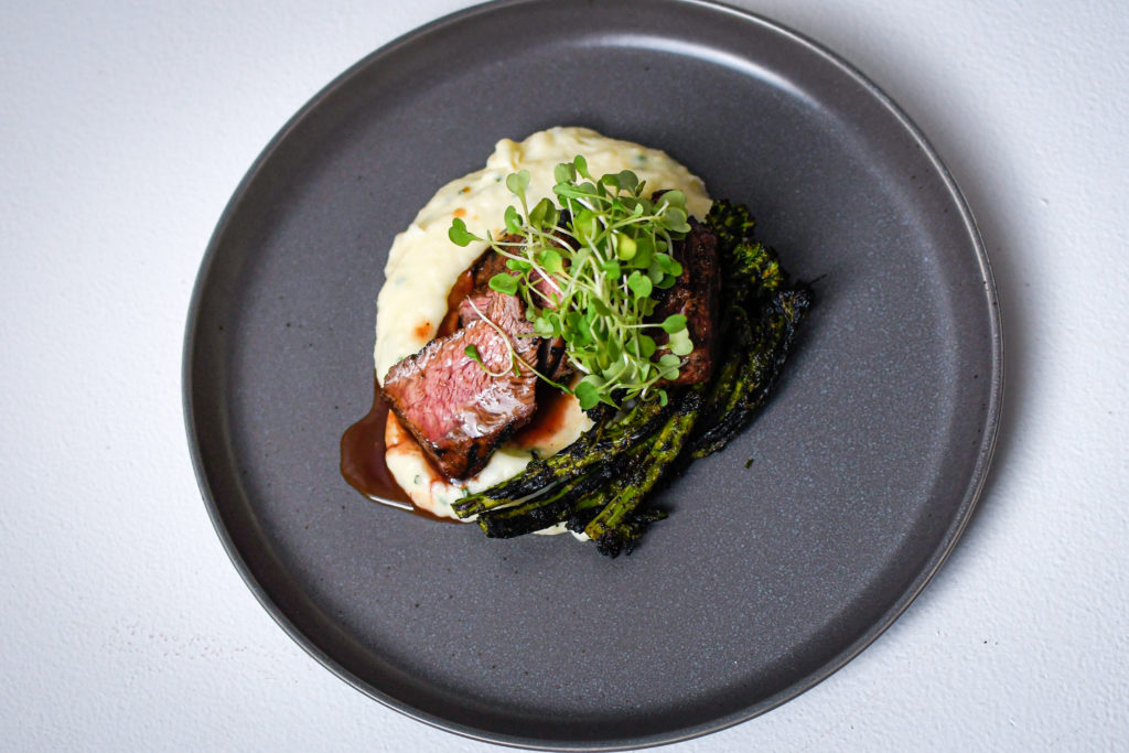 Slices of Adobe Flatiron Steak sit of a bed of mashed potatoes with charred broccolini on the side. It's topped with microgreens. This sits of a grey/black plate on a white textured background.