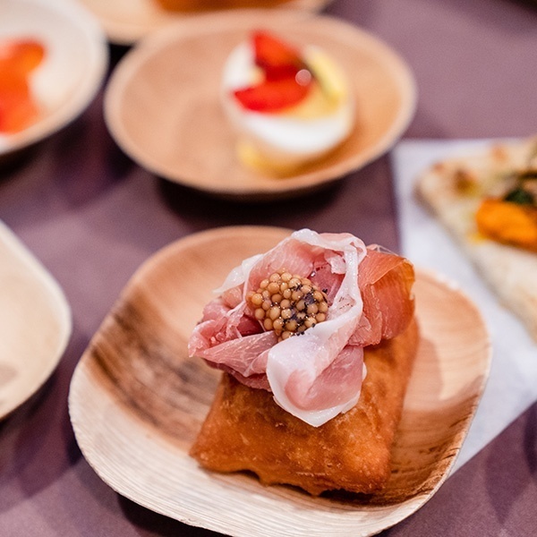 Close up of multiple small plates on veterra plates. The center on in focus is a piece a beignet topped with shaved prosciutto and pickled mustard seed. The rest of the plates surrounding it are slightly out of focus and incluses a piece of flatbread and deviled eggs with beet cured trout