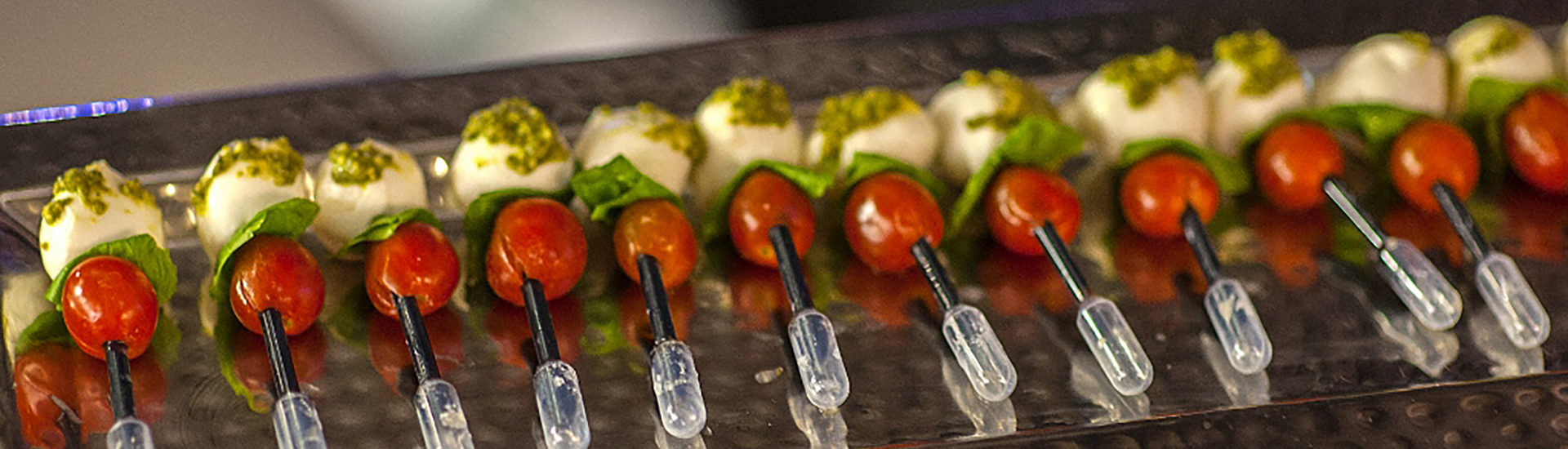Multiple hors d'oeuvres of a piece of mozzarella ball with pesto on top, a basil leaf, cherry tomato on a plastic pipette full of balsamic vinegar.