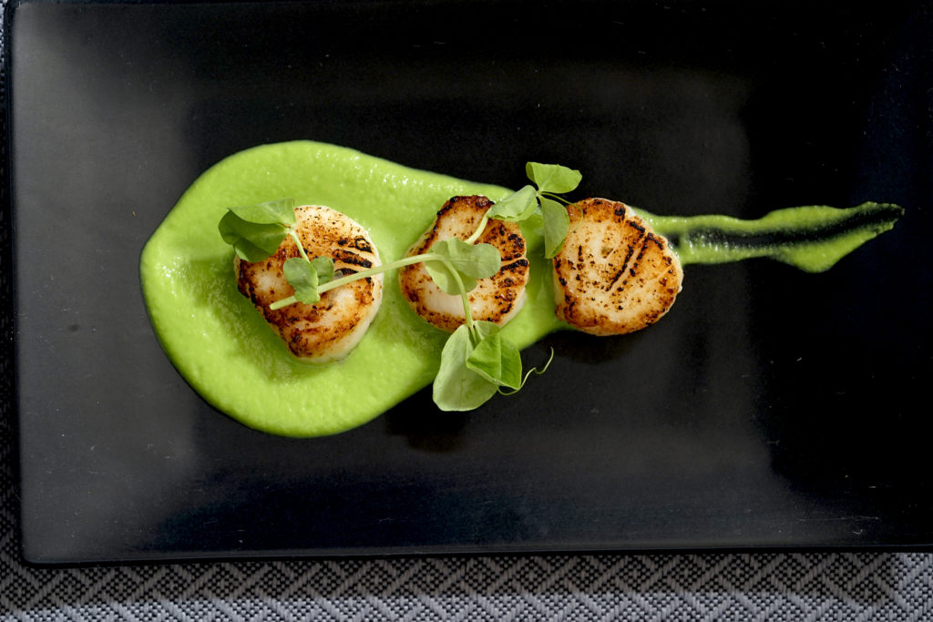 3 seared scalloped siting on a swoosh of pea puree with a piece of herb on top on a black rectangle plate.
