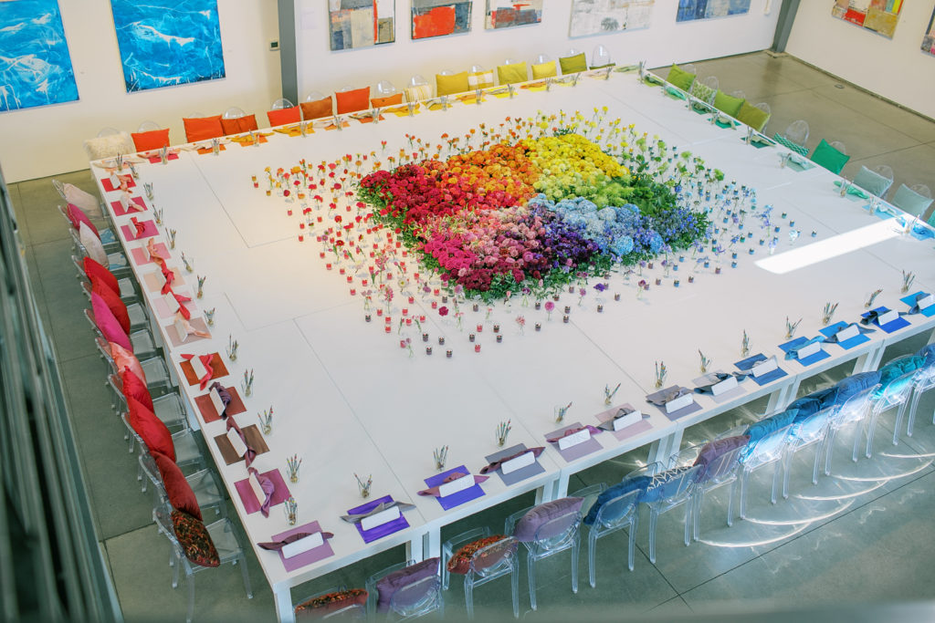 Above shot of a giant white white made from pushing multiple tables together with a colorful flower display in the center in an ombre rainbow surround by flowers in individual glasses and candles. Around the edges of the table are colorful seating with a pillow, place mat and napkin in a rainbow.