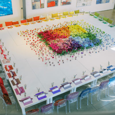 Above shot of a giant white white made from pushing multiple tables together with a colorful flower display in the center in an ombre rainbow surround by flowers in individual glasses and candles. Around the edges of the table are colorful seating with a pillow, place mat and napkin in a rainbow.