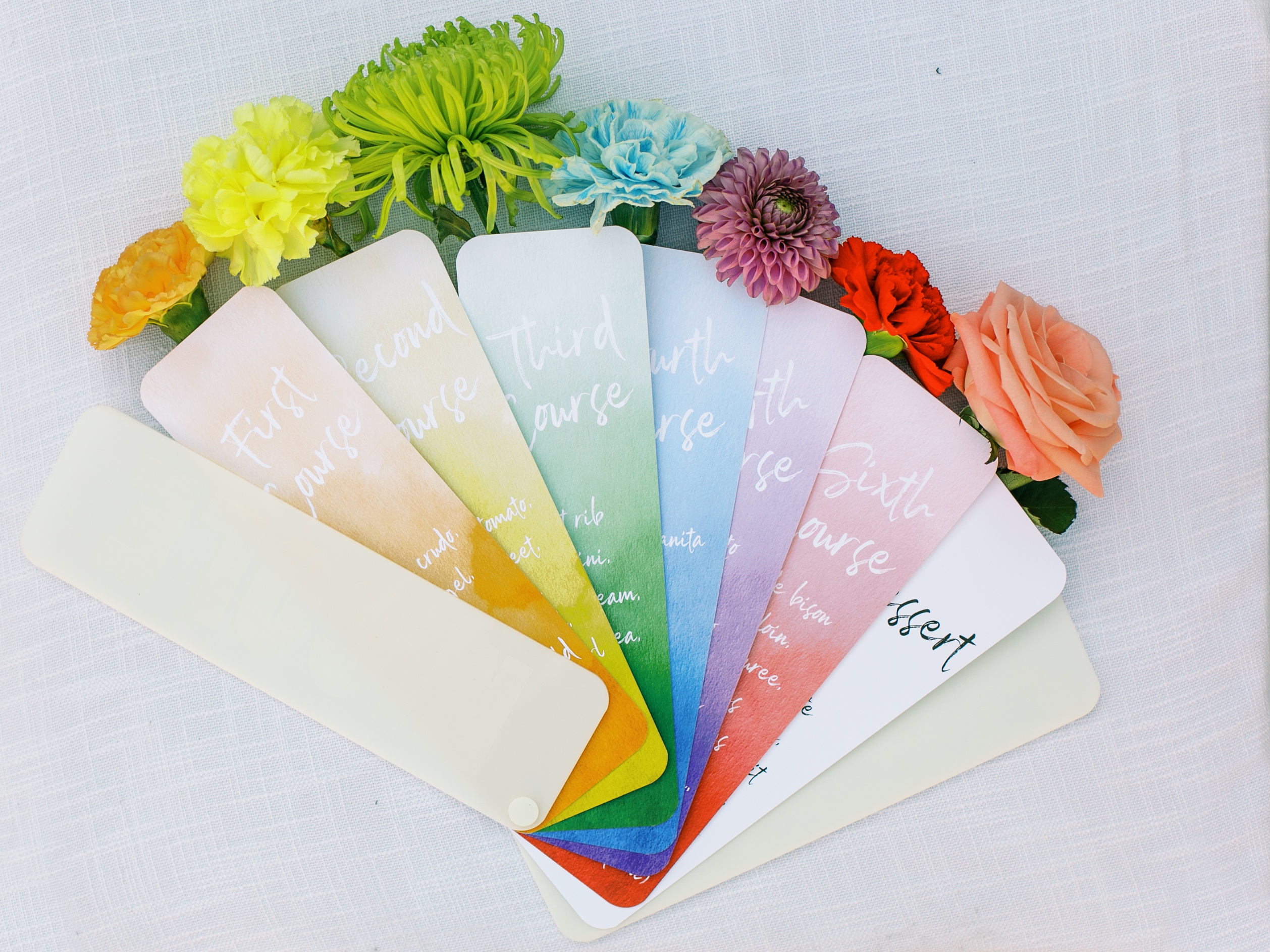 Thin rectangle menu cards are individually ombre with the darkest of the color at the bottom and getting lighter to the top. The cards have white text in a rainbow starting with orange and ending with red. Behind each card is a single flower that colors matches the card.