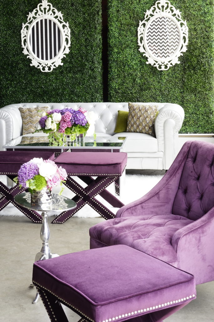 In the background are 2 hedge walls with mirror frames on them. In front of the walls are a white couch with gold and silver sequin pillows. In front of the couch are a glass pub table with purple white and green floral centerpiece. In the front is a purple chair with a purple square stool with silver studs.