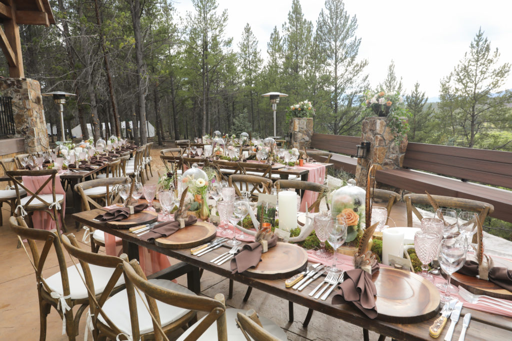 Four rustic wooden tables outsidw on a patio with wooden chairs with white cushions. On the tables are wooden circle used as placemats with a brown napkin on top with some feathers as decorations. Silver silverware with a pink table runner. It's decorated with green moss, pink chalice glasses, clear wine glasses, candles, twinkle slights and roses with a cloche on top. There are lots of trees with the background.