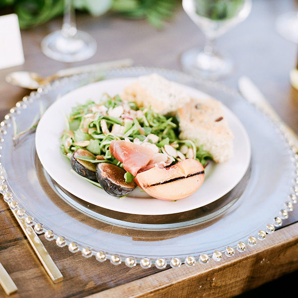 Grilled Peaches and Fig Salad with prosciutto, arugula and sliced almonds and 2 pieces of bread on a whtie plate on a clear charger with small balls surrounding it on a wooden table with gold silverware