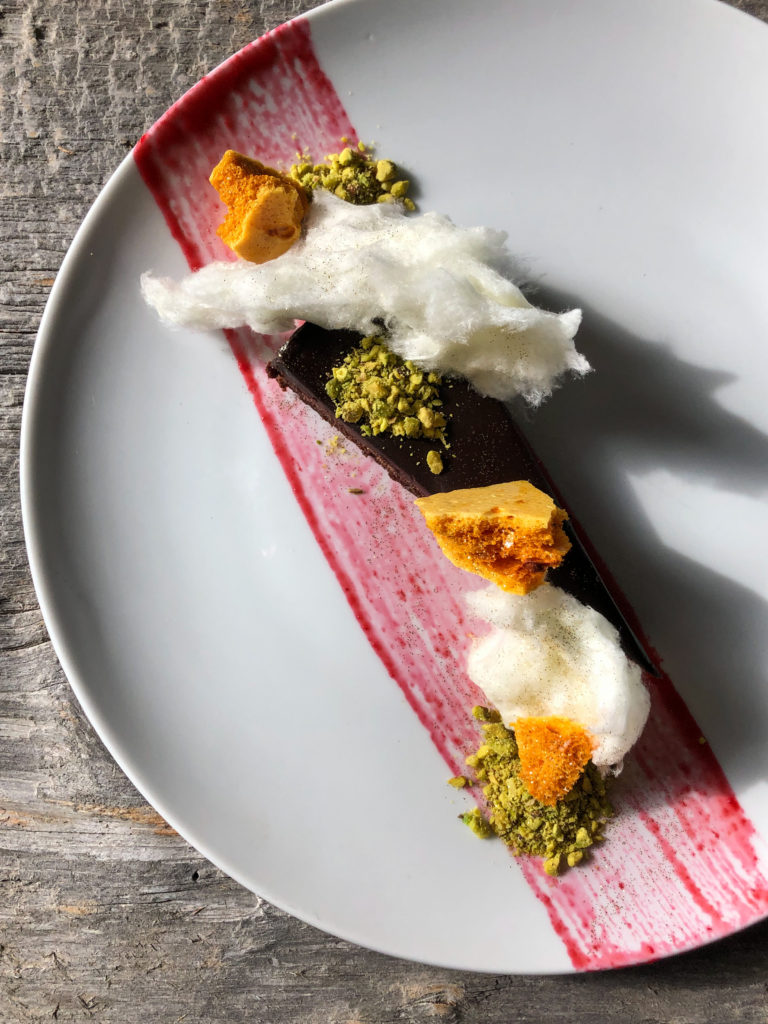 A piece of flourless chocolate cake plated with pieces of honey comb, cotton candy and crushed pistachios. It has red sauce swooshed on a white plate underneath a wood background.