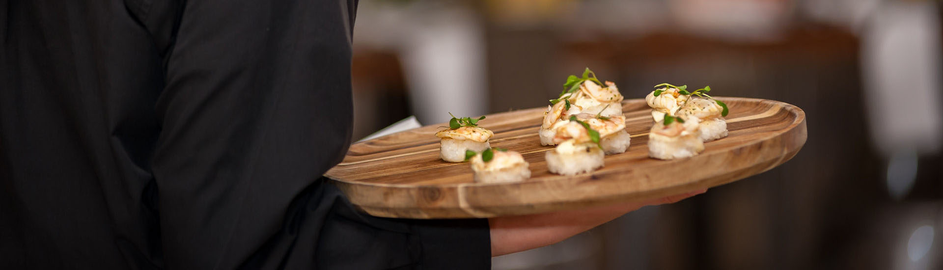Close up of a server in a black uniform holding a circle wooden passing tray which has nigiri on it with a little bit of microgreens on top
